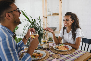 Young cheerful multi-ethnic couple eating pasta, drinking vine having lunch together at home