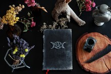 Flat Lay Of Witch's Altar Space With Hand Made Old Looking Book Grimoire And Other Various Items - Dried Flowers Nature Elements, Candles, Ink Bottle With Black Feather Next To It On Dark Black Table