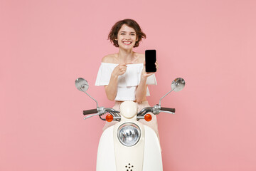 Smiling young woman in white summer clothes pointing index finger on mobile phone with blank empty screen mock up copy space driving moped isolated on pastel pink colour background studio portrait.