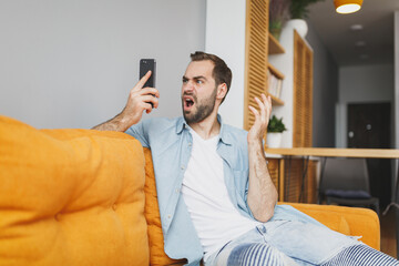 Canvas Print - Shocked irritated displeased young bearded man 20s wearing casual white t-shirt blue shirt spreading hands talking on mobile cell phone sitting on couch resting spending time in living room at home.