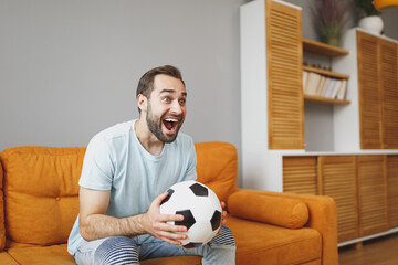 Wall Mural - Excited young bearded man football fan 20s wearing basic blue t-shirt cheer up support favorite team with soccer ball keeping mouth open sitting on couch resting spending time in living room at home.