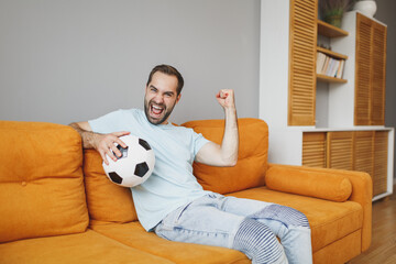 Canvas Print - Joyful young bearded man football fan 20s in basic blue t-shirt cheer up support favorite team with soccer ball doing winner gesture sitting on couch resting spending time in living room at home.