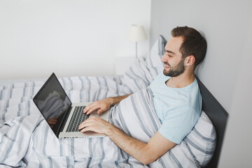 Side view of smiling young bearded man 20s wearing basic blue t-shirt working on laptop pc computer lying in bed with striped sheet pillow blanket resting relaxing spending time in bedroom at home.