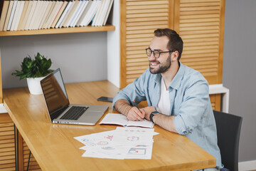 Canvas Print - Handsome smiling attractive young bearded business man 20s wearing blue shirt glasses sitting at desk with papers document working on laptop pc computer writing in notebook at home or office.