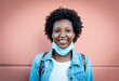 beautiful smile of a black girl, wearing a protective and medical mask lowered under her chin, face expressing positive and optimistic thinking, pastel coral pink background