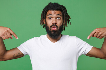 Wall Mural - Shocked amazed young african american man guy with dreadlocks 20s in white casual t-shirt posing pointing index fingers down on mock up copy space isolated on green color background studio portrait.
