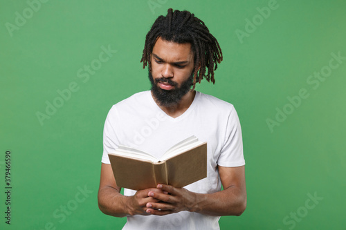 Fotografía Pensive concentrated young african american man guy with dreadlocks 20s wearing white casual t-shirt posing holding in hands reading book studying isolated on green color background studio portrait