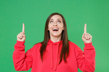 Wall Mural - Excited surprised young brunette woman 20s wearing bright red casual streetwear hoodie posing pointing index fingers up on mock up copy space isolated on green color wall background studio portrait.