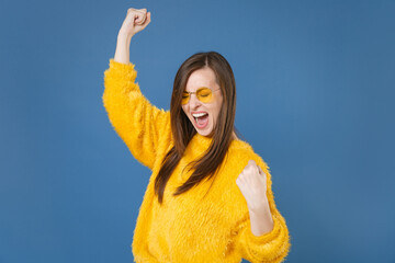 Wall Mural - Happy joyful screaming young brunette woman 20s wearing yellow fur sweater eyeglasses posing clenching fists doing winner gesture keeping eyes closed isolated on blue color background studio portrait.