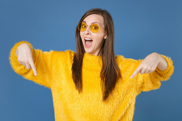 Wall Mural - Shocked surprised young brunette woman 20s in yellow fur sweater eyeglasses posing pointing index fingers down on mock up copy space looking camera isolated on blue color background studio portrait.