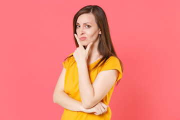 Wall Mural - Pensive confused perplexed concerned young brunette woman 20s wearing yellow casual t-shirt posing standing put hand prop up on chin looking camera isolated on pink color background studio portrait.