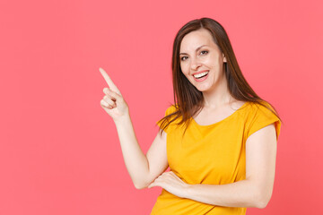 Wall Mural - Cheerful smiling beautiful young brunette woman 20s wearing yellow casual t-shirt posing pointing index finger aside up on mock up copy space isolated on pink color wall background studio portrait.