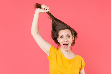 Excited surprised shocked young brunette woman 20s in yellow casual t-shirt posing holding hair like ponytail fooling around keeping mouth open isolated on pink color wall background studio portrait.