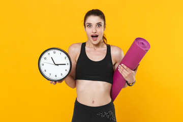 Wall Mural - Excited shocked young fitness sporty woman 20s wearing black sportswear posing training working out hold clock yoga mat keeping mouth open isolated on bright yellow color background studio portrait.