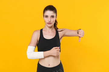 Wall Mural - Dissatisfied young fitness sporty woman 20s wearing black sportswear posing training get injured arm elbow with elastic bandage showing thumb down isolated on bright yellow background studio portrait.