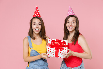 Wall Mural - Two excited cheerful young women friends 20s in casual denim clothes birthday hats celebrating hold red present box with gift ribbon bow isolated on pastel pink colour background, studio portrait.