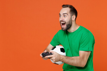 Wall Mural - Excited young man football fan in green t-shirt cheer up support favorite team with soccer ball play pc game with joystick console isolated on orange background studio. People sport leisure concept.