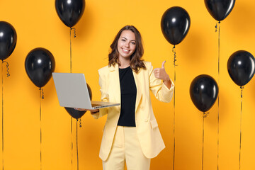 Smiling cheerful young brunette woman 20s wearing basic light suit jacket using laptop pc computer showing thumb up on yellow colour background with air balloons studio portrait. Black friday sale.