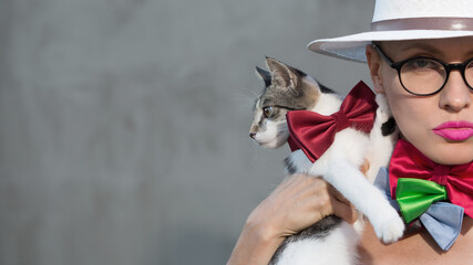 Wall Mural - Portrait of a beautiful young woman wearing hat, glasses, many bow ties and holding a cat with a tie, copy space