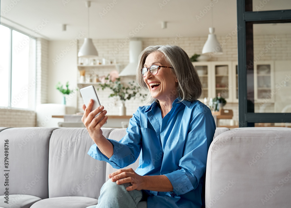 Happy mature old 60s woman holding smartphone using mobile phone app for video call, laughing while watching funny video, feeling excited winning online lottery bid on cellphone sits on couch at home. - obrazy, fototapety, plakaty 