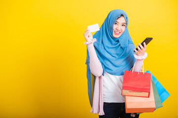 Asian Muslim Arab, Portrait of happy beautiful young woman Islam religious wear veil hijab funny smile she using smartphone on hand and hold shopping bags, studio shot isolated on yellow background