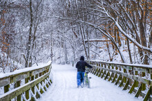 Teenage Boy Walking On Wooden Bridge Covered With Deep Snow On Snowy Day In Midwest And Pulling His Bicycle With Him