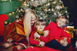 © hetmanstock2 - Beautiful mother in a red dress. Family sitting near christmas gifts. Little girl and boy near christmas tree