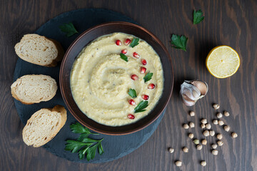 Canvas Print - Top view of savory hummus made of chickpeas with tahini, lemon juice and garlic decorated with pomegranate seeds and parsley on tray with bread slices on wooden background at kitchen. Horizontal