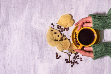 Female hands in a warm sweater hold a cup of coffee on a light rustic wooden table background with autumn leaves and coffee grains