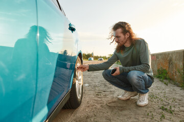 Wall Mural - Young caucasian man checking air pressure of car tire on local road side while traveling, Male driver having troubles with his auto, checking wheel after car breakdown