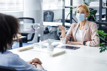 Wall Mural - African american lady speaks to business woman in protective mask through glass partition in office interior