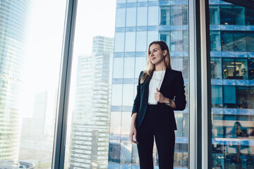 Wall Mural - Cheerful blonde female manager in formal outfit enjoying job and successful career in business, smiling beautiful woman boss dressed in elegant suit standing near panoramic windows in office
