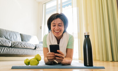 Beautiful brunette older woman training gym at home holding smartphone. Senior female workout with fitness app on the phone. Concept about senior people and sport.