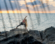 Young Caucasian Woman With Long Blond Hair In Yoga Clothes Doing Yoga Asana On The Rocks By The Sea At Colorful Dramatic Sunrise On  Tropical Island. View Through Palm Leaves.