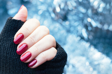 Wall Mural - Female hand in black knitted gloves mitts with a beautiful glossy manicure - burgundy, dark red, cherry color nails on background of blue Christmas tinsel garland. Selective focus. Blurred background