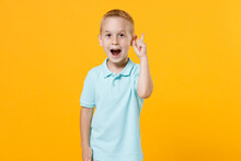 Smiling Little Fun Male Kid Boy 5-6 Years Old Wearing Stylish Blue Turquoise T-shirt Polo Holding Index Finger Up With Great New Idea Isolated On Yellow Color Wall Background, Child Studio Portrait.