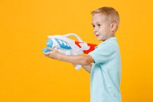Smiling Little Fun Male Fair-haired Brown-eyed Kid Boy 5-6 Years Old Wearing Stylish Blue Turquoise T-shirt Polo Hold In Hand Toy Water Gun Isolated On Yellow Color Background, Child Studio Portrait.
