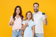 © ViDi Studio - Smiling young parents mom dad with child kid daughter teen girl in basic t-shirts hold credit bank card showing thumb up isolated on yellow background studio portrait. Family day parenthood concept.