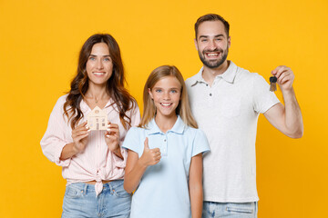 Wall Mural - Smiling young parents mom dad with child kid daughter teen girl in basic t-shirts hold house bunch of apartment keys showing thumb up isolated on yellow background studio portrait. Family day concept.