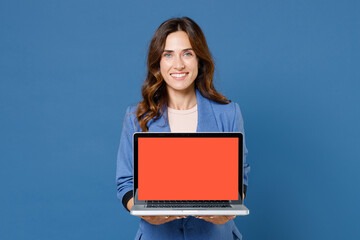 Smiling young brunette woman 20s wearing basic jacket hold laptop pc computer with blank empty screen mock up copy space looking camera isolated on bright blue colour wall background studio portrait.