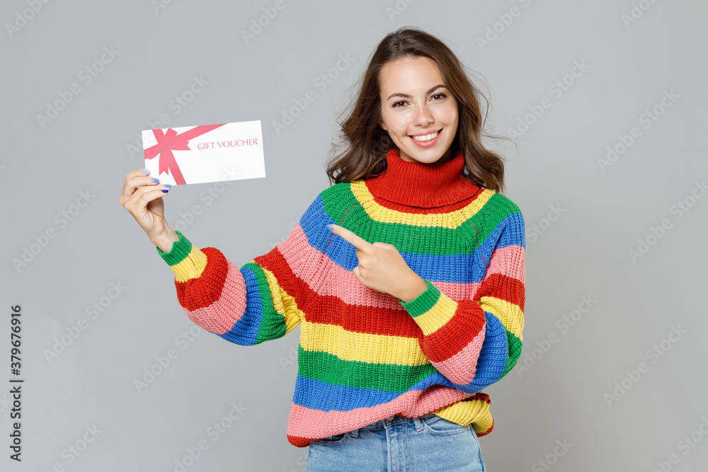 Smiling cheerful young brunette woman 20s wearing casual colorful knitted sweater standing pointing index finger on gift certificate looking camera isolated on grey colour background, studio portrait. - obrazy, fototapety, plakaty 