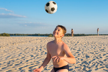 Happy boy cheerfully plays football on the beach, on a sunny day. Outdoor sports games. Active healthy lifestyle concept.