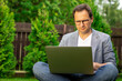 © Model Republic - Focused unshaven mature European 40s businessman in glasses working outdoors sitting with crossed legs on green grass with laptop on blurred green natural background. writer writes novel.