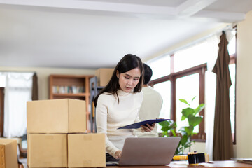 Startup small business. Young asian woman holding document paper and using computer laptop for checking product in the parcels. Young man packing goods for delivery to customer