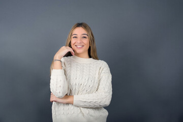 Beautiful blonde young woman wearing white sweater feeling confident against gray studio background.