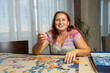 © Andres - Beautiful woman with brown hair and in a colorful dress doing puzzle in the living room at home