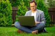 © Model Republic - Adult smart businessman in glasses working on laptop outdoors, sitting on grass, sell cryptocurrency, buing up shares, check stock market, online trading, e-commerce, financial management concept