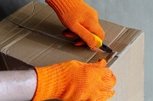 A Worker Wearing Protective Gloves Opens A Cardboard Box.