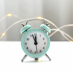 A small blue alarm clock stands on a table with garland lights. Holiday concept for Christmas and New Year.