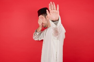 Wall Mural - young caucasian muslim man wearing djellaba and traditional hat over red background covers eyes with palm and doing stop gesture, tries to hide from everybody. Don't look at me, I don't want to see,